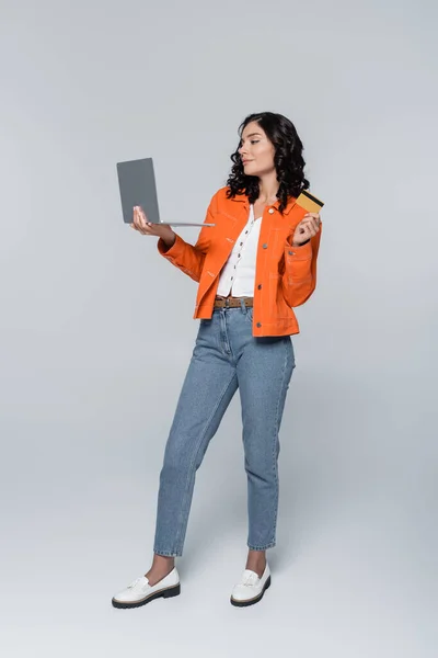 Comprimento total da jovem mulher em jaqueta laranja segurando laptop e cartão de crédito no cinza — Fotografia de Stock