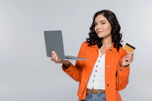 Задоволена молода жінка в помаранчевій куртці тримає ноутбук і кредитну картку ізольовано на сірому — стокове фото
