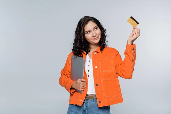 Pleased woman in orange jacket holding laptop and looking at credit card isolated on grey — Stock Photo