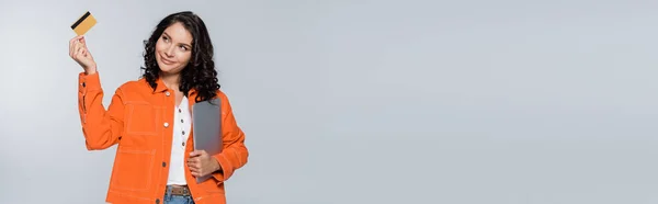 Mujer complacida en chaqueta naranja sosteniendo el ordenador portátil y mirando la tarjeta de crédito aislada en gris, pancarta - foto de stock