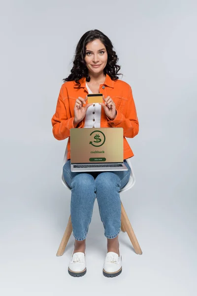 Young woman in orange jacket holding credit card near laptop with cashback lettering on screen on grey — Stock Photo