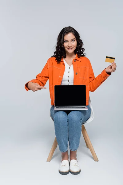 Cheerful young woman in orange jacket pointing at laptop with blank screen and holding credit card on grey — Stock Photo