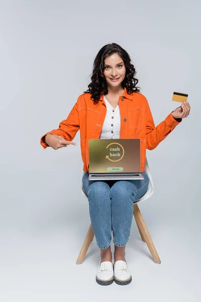 Fröhliche junge Frau in orangefarbener Jacke zeigt auf Laptop mit Bargeld auf dem Bildschirm und hält Kreditkarte auf grau — Stockfoto