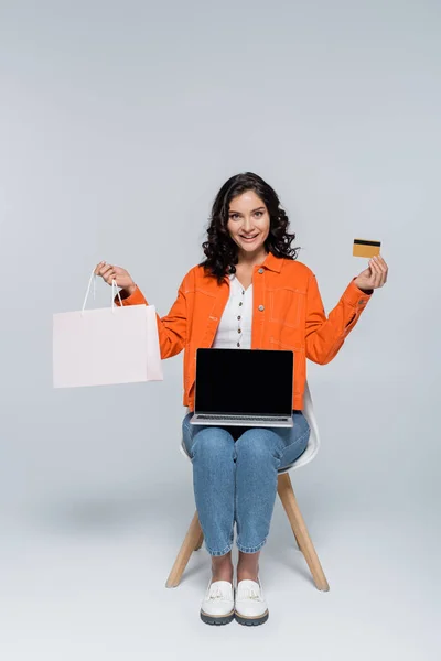 Mujer alegre sosteniendo bolsa de compras y tarjeta de crédito cerca del ordenador portátil con pantalla en blanco en gris - foto de stock