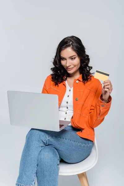 Mujer joven y complacida con chaqueta naranja usando portátil y la celebración de la tarjeta de crédito aislado en gris - foto de stock