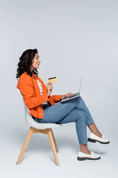 Vue latérale de la jeune femme en veste orange à l'aide d'un ordinateur portable et tenant la carte de crédit sur gris — Photo de stock