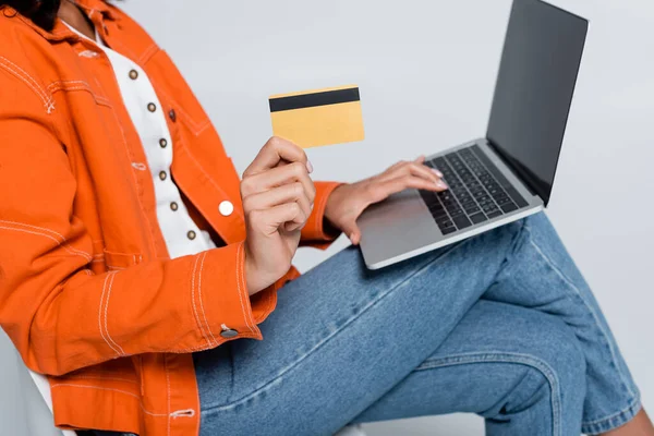 Cropped view of woman in orange jacket using laptop and holding credit card isolated on grey — Stock Photo