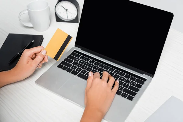 Cropped view of woman holding credit card near laptop with blank screen and notebook on white desk — Stock Photo