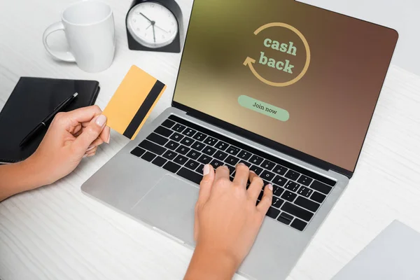 Ausgeschnittene Ansicht einer Frau mit Kreditkarte in der Nähe von Laptop mit Bargeld auf dem Bildschirm und Notizbuch auf weißem Schreibtisch — Stockfoto