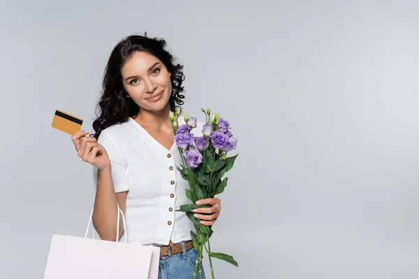 Mujer complacida sosteniendo ramo de flores, bolsa de compras y tarjeta de crédito aislada en gris - foto de stock