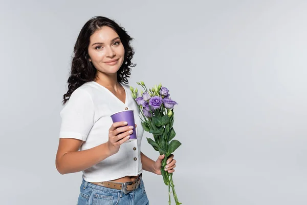 Pleased young woman holding bouquet of purple flowers and paper cup isolated on grey — Stock Photo