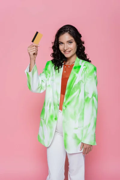 Pleased young woman in tie-dye blazer holding credit card isolated on pink — Stock Photo