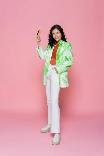 Pleine longueur de jeune femme heureuse en blazer tie-dye tenant la carte de crédit et posant avec la main dans la poche sur rose — Photo de stock