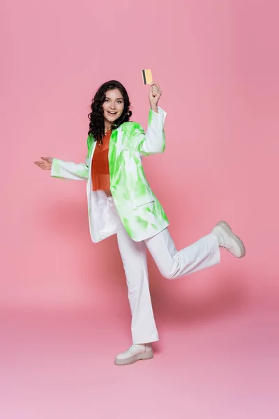 Comprimento total de feliz jovem mulher em tie-dye blazer segurando cartão de crédito em rosa — Fotografia de Stock