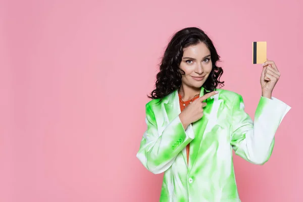 Cheerful young woman in tie-dye blazer pointing at credit card isolated on pink — Stock Photo