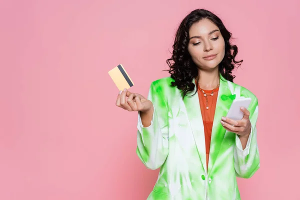 Mujer joven en blazer de tinte de corbata con tarjeta de crédito y teléfono inteligente aislado en rosa - foto de stock