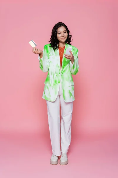 Longitud completa de alegre mujer joven en corbata chaqueta de tinte con tarjeta de crédito y teléfono inteligente en rosa - foto de stock
