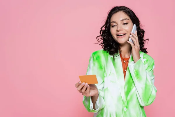 Feliz joven mujer en corbata blazer tinte celebración de la tarjeta de crédito y hablar en el teléfono inteligente aislado en rosa - foto de stock