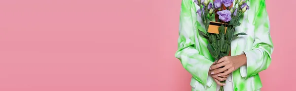 Vista recortada de mujer joven en corbata chaqueta de tinte con flores de color púrpura con tarjeta de crédito aislado en rosa, bandera — Stock Photo