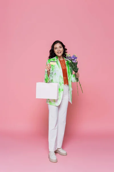 Full length of smiling young woman in tie dye blazer holding credit card, shopping bag and flowers on pink — Stock Photo