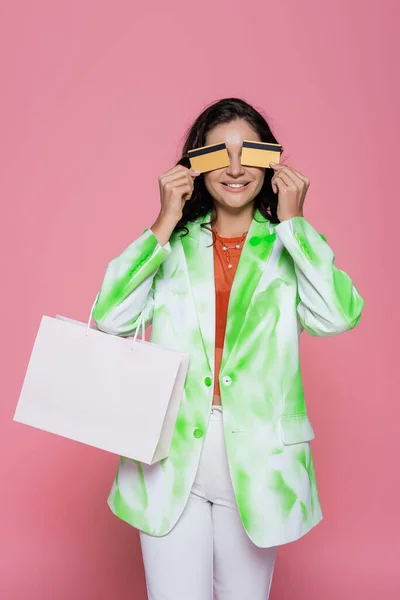 Jeune femme positive en blazer de teinture cravate couvrant les yeux avec des cartes de crédit et tenant sac à provisions isolé sur rose — Photo de stock