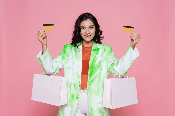 Alegre joven mujer en corbata blazer tinte celebración de tarjetas de crédito y bolsas de compras aislados en rosa - foto de stock
