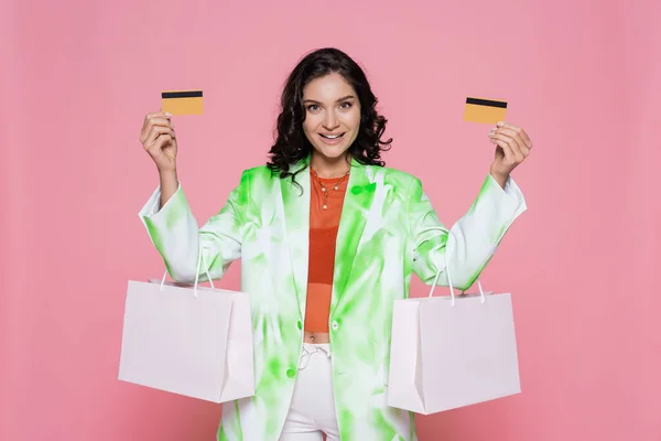 Cheerful young woman in tie dye blazer holding credit cards and shopping bags isolated on pink — Stock Photo
