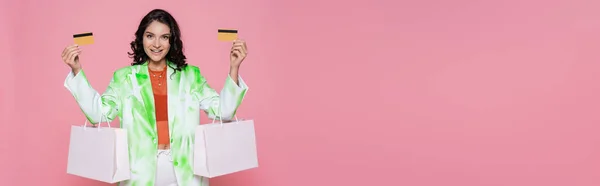 Cheerful young woman in tie dye blazer holding credit cards and shopping bags isolated on pink, banner — Stock Photo