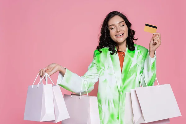 Alegre joven mujer en corbata chaqueta de tinte con tarjeta de crédito y bolsas de compras aislados en rosa - foto de stock