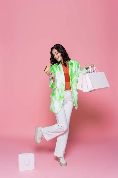 Longitud completa de alegre mujer joven en corbata chaqueta de tinte que sostiene la tarjeta de crédito y bolsas de compras en rosa - foto de stock