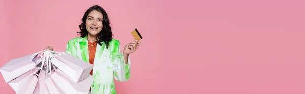 Joyeuse jeune femme en blazer de teinture cravate tenant carte de crédit et sacs à provisions isolés sur rose, bannière — Photo de stock