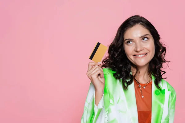 Sognante giovane donna in cravatta colorante blazer in possesso di carta di credito e sorridente isolato su rosa — Foto stock