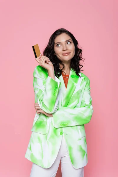 Femme rêveuse en veste de teinture cravate verte tenant carte de crédit et souriant isolé sur rose — Photo de stock