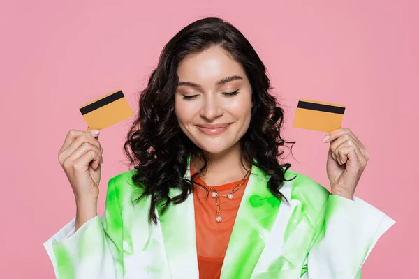 Pleased woman in green tie dye blazer holding credit cards and smiling isolated on pink — Stock Photo