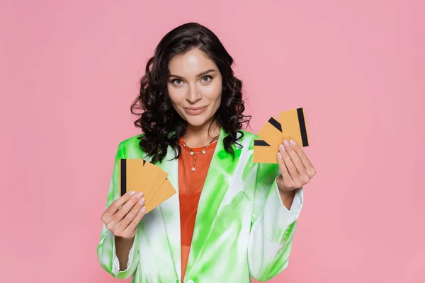 Mujer positiva en verde corbata blazer tinte celebración de tarjetas de crédito y sonriendo aislado en rosa - foto de stock