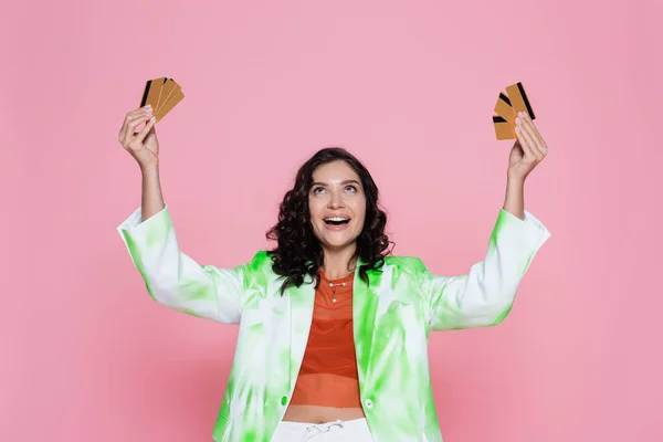 Erstaunt junge Frau im grünen Krawattenfärbeblazer schaut auf Kreditkarten und lächelt isoliert auf rosa — Stockfoto