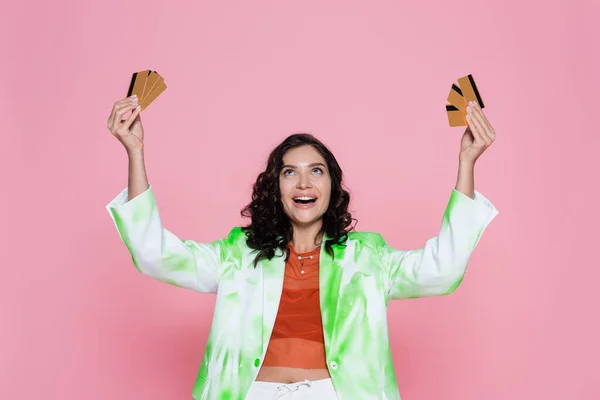 Mujer joven asombrada en verde corbata chaqueta de tinte mirando las tarjetas de crédito y sonriendo aislado en rosa - foto de stock