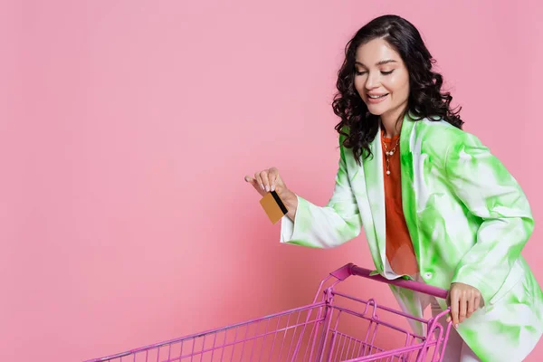 Mujer alegre en blazer verde con tarjeta de crédito y de pie cerca del carrito de compras en rosa - foto de stock