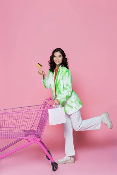 Pleine longueur de femme joyeuse en veste verte tenant carte de crédit et sac en papier près du panier sur rose — Photo de stock