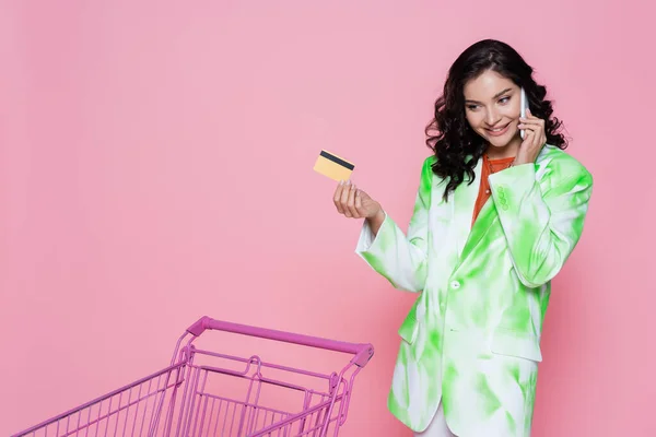 Cheerful woman in green blazer holding credit card and talking on smartphone near shopping cart isolated on pink — Stock Photo