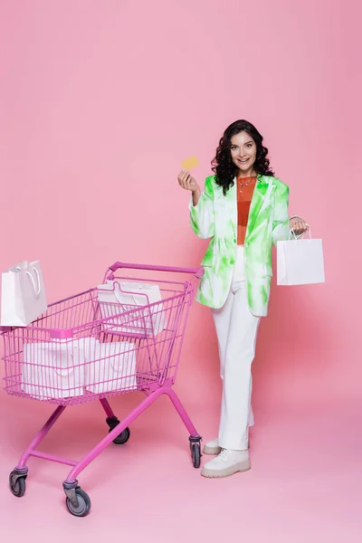 Pleine longueur de femme joyeuse en veste verte tenant la carte de crédit près du panier avec des sacs en papier sur rose — Photo de stock