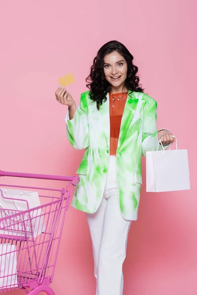 Mujer alegre en blazer verde con tarjeta de crédito cerca del carrito de la compra con bolsas de papel en rosa - foto de stock