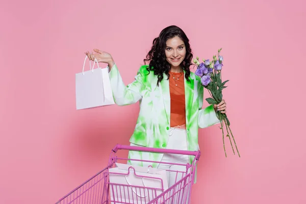 Cheerful young woman in green blazer holding flowers near shopping cart with paper bags on pink — Stock Photo