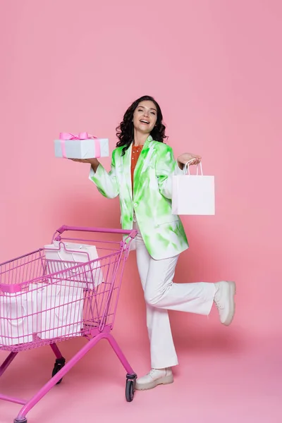 Volle Länge der fröhlichen Frau im Blazer hält verpackten Geschenkkarton in der Nähe von Einkaufswagen mit Papiertüten auf rosa — Stockfoto