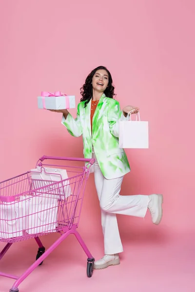 Pleine longueur de femme gaie dans le blazer tenant boîte cadeau enveloppée près du panier avec des sacs en papier sur rose — Photo de stock