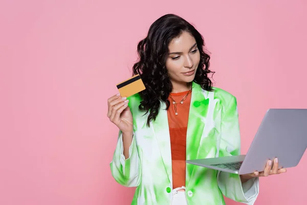 Morena mujer en blazer celebración de la tarjeta de crédito y portátil aislado en rosa - foto de stock
