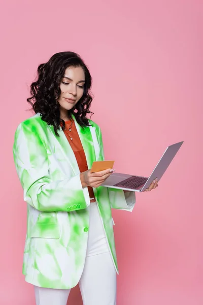 Joven morena mujer en blazer celebración de tarjeta de crédito y portátil aislado en rosa - foto de stock