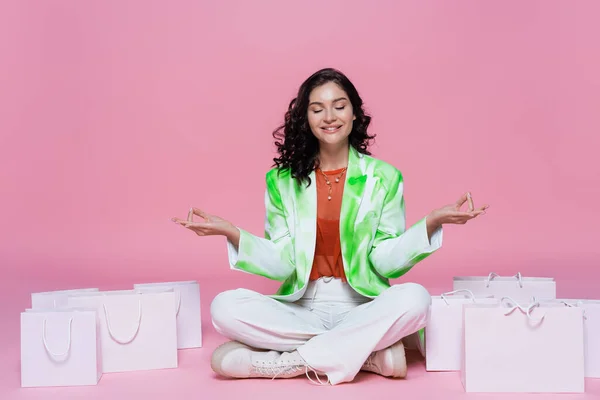 Mujer morena feliz en blazer sentado en pose de yoga mientras medita cerca de bolsas de compras en rosa - foto de stock