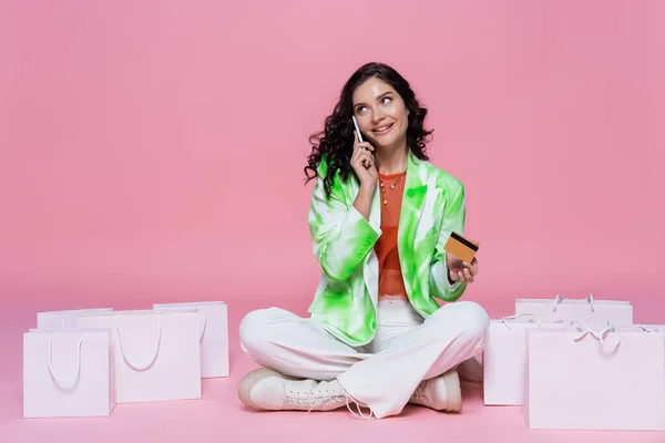 Mujer feliz en blazer hablando en el teléfono inteligente mientras sostiene la tarjeta de crédito cerca de bolsas de compras en rosa - foto de stock