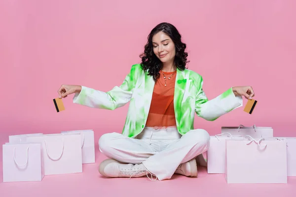 Femme heureuse en veste verte tenant des cartes de crédit tout en étant assis avec les jambes croisées près des sacs à provisions sur rose — Photo de stock
